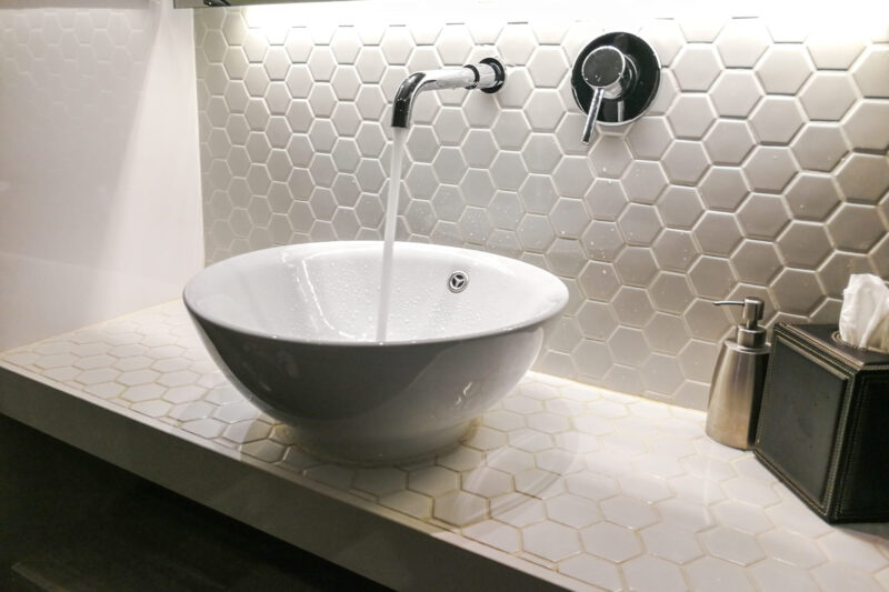Voda iz zidne slavine teče u okrugli lavabo na ploči sa belim šestougaonim pločicama